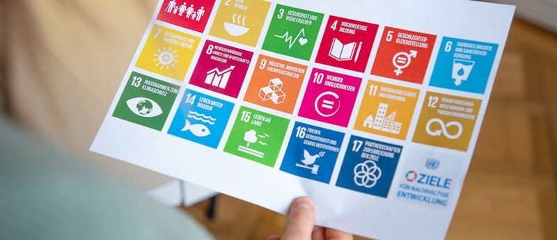 Eine Hand hält ein Blatt mit einer Abbildung der SDGs, farbige Kacheln, hoch.